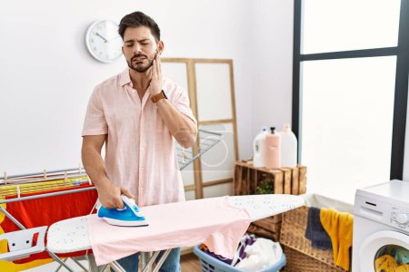 Foto de Young man with beard ironing clothes at home touching mouth with hand with painful expression because of toothache or dental illness on teeth. dentist concept. - Imagen libre de derechos