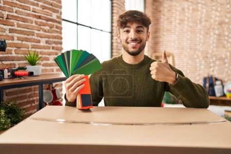 Foto de Arab man with beard moving to a new home choosing walls paint smiling happy and positive, thumb up doing excellent and approval sign - Imagen libre de derechos
