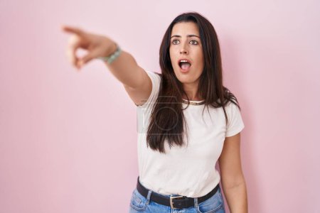 Téléchargez les photos : Jeune femme brune debout sur fond rose pointant du doigt surpris devant, bouche ouverte expression étonnante, quelque chose sur le devant - en image libre de droit
