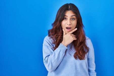 Téléchargez les photos : Hispanic young woman standing over blue background looking fascinated with disbelief, surprise and amazed expression with hands on chin - en image libre de droit