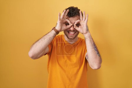Foto de Un joven hispano de pie sobre un fondo amarillo haciendo un buen gesto como binoculares sacando la lengua, ojos mirando a través de los dedos. expresión loca. - Imagen libre de derechos
