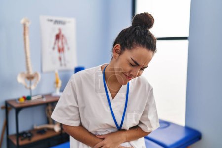 Foto de Young hispanic woman working at rehabilitation clinic with hand on stomach because nausea, painful disease feeling unwell. ache concept. - Imagen libre de derechos