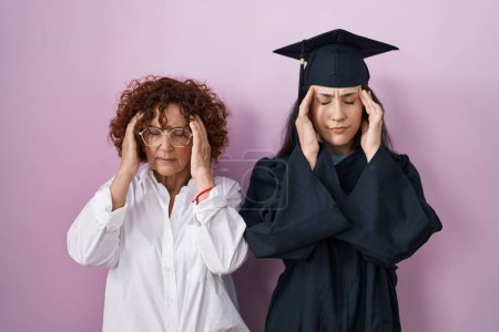 Foto de Madre e hija hispanas usando gorra de graduación y bata de ceremonia con la mano en la cabeza para el dolor en la cabeza porque el estrés. padecer migraña. - Imagen libre de derechos