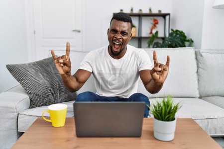 Téléchargez les photos : Jeune homme africain utilisant un ordinateur portable à la maison criant avec une expression folle faire symbole rock avec les mains en l'air. star de la musique. concept lourd. - en image libre de droit