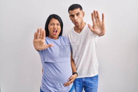 Photo for Young hispanic couple expecting a baby standing over background doing stop gesture with hands palms, angry and frustration expression - Royalty Free Image