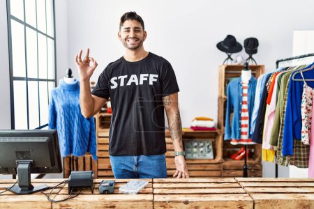 Foto de Joven hombre hispano que trabaja en la boutique minorista sonriendo positiva haciendo signo ok con la mano y los dedos. expresión exitosa. - Imagen libre de derechos