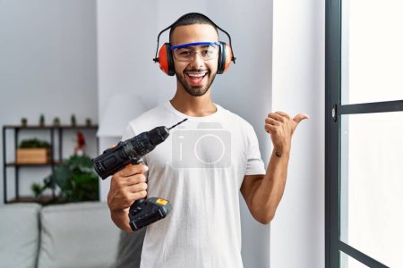 Téléchargez les photos : African american man holding screwdriver wearing ear protection at home pointing thumb up to the side smiling happy with open mouth - en image libre de droit