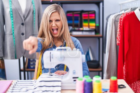 Photo for Blonde woman dressmaker designer using sew machine pointing displeased and frustrated to the camera, angry and furious with you - Royalty Free Image