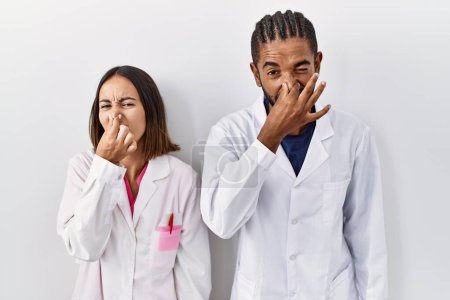 Téléchargez les photos : Jeunes médecins hispaniques debout sur fond blanc sentant quelque chose de puant et dégoûtant, odeur intolérable, retenant souffle avec les doigts sur le nez. mauvaise odeur - en image libre de droit