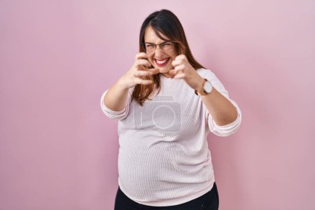 Photo for Pregnant woman standing over pink background shouting frustrated with rage, hands trying to strangle, yelling mad - Royalty Free Image