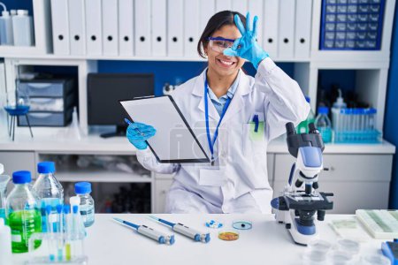 Foto de Hispanic young woman working at scientist laboratory doing ok gesture with hand smiling, eye looking through fingers with happy face. - Imagen libre de derechos