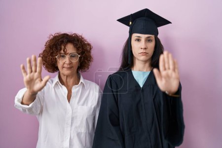 Photo for Hispanic mother and daughter wearing graduation cap and ceremony robe doing stop sing with palm of the hand. warning expression with negative and serious gesture on the face. - Royalty Free Image