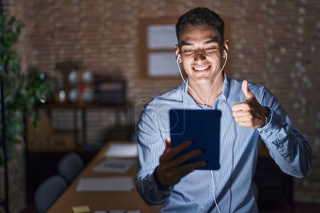 Photo for Handsome hispanic man working at the office at night doing happy thumbs up gesture with hand. approving expression looking at the camera showing success. - Royalty Free Image