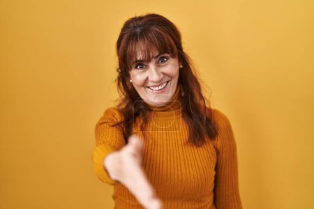 Foto de Mujer hispana de mediana edad de pie sobre fondo amarillo sonriente amistoso ofreciendo apretón de manos como saludo y bienvenida. negocios exitosos. - Imagen libre de derechos