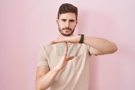 Foto de Hombre hispano con barba de pie sobre fondo rosa haciendo un gesto de tiempo fuera con las manos, rostro frustrado y serio - Imagen libre de derechos