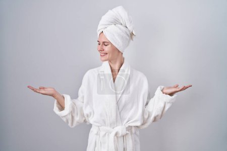 Foto de Mujer rubia caucásica vistiendo albornoz sonriente mostrando ambas manos palmas abiertas, presentando y comparando publicidad y equilibrio - Imagen libre de derechos