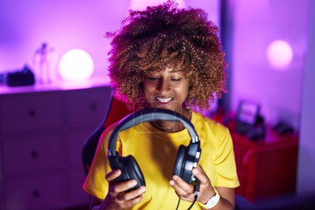 Téléchargez les photos : Femme afro-américaine streamer souriant confiant tenant écouteurs à la salle de jeux - en image libre de droit