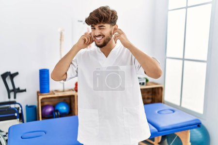 Photo for Young arab man working at pain recovery clinic covering ears with fingers with annoyed expression for the noise of loud music. deaf concept. - Royalty Free Image