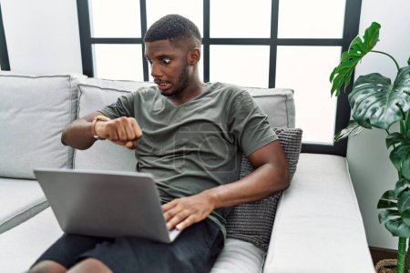 Foto de Young african american man using laptop at home sitting on the sofa looking at the watch time worried, afraid of getting late - Imagen libre de derechos