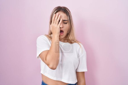Foto de Young blonde woman standing over pink background yawning tired covering half face, eye and mouth with hand. face hurts in pain. - Imagen libre de derechos