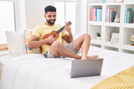 Téléchargez les photos : Jeune homme arabe ayant une leçon d'ukulélé en ligne assis sur le lit dans la chambre - en image libre de droit