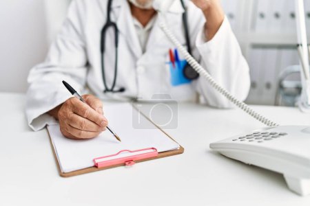Téléchargez les photos : Homme âgé aux cheveux gris portant l'uniforme de médecin parlant au téléphone à la clinique - en image libre de droit