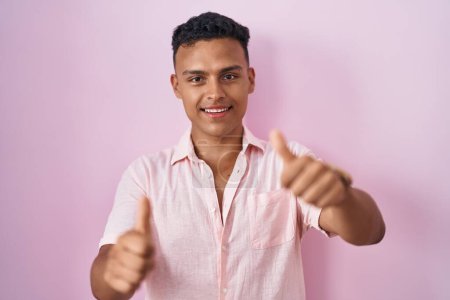 Téléchargez les photos : Jeune homme hispanique debout sur fond rose approuvant faire un geste positif avec la main, pouces levés souriant et heureux pour le succès. geste gagnant. - en image libre de droit