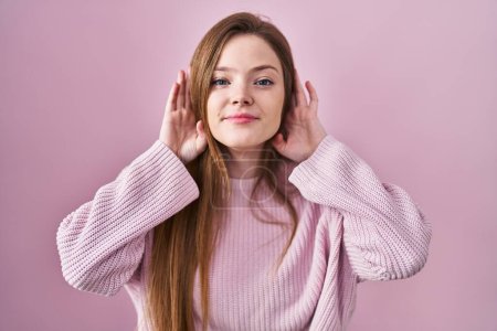 Foto de Mujer caucásica joven de pie sobre fondo rosa tratando de escuchar ambas manos en el gesto de la oreja, curioso por chismes. problema auditivo, sordo - Imagen libre de derechos