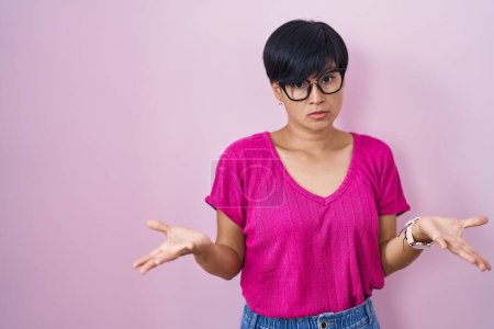 Foto de Joven mujer asiática con pelo corto de pie sobre fondo rosa despistado y confundido con los brazos abiertos, sin concepto de idea. - Imagen libre de derechos