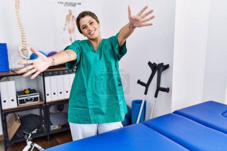 Foto de Mujer hispana joven vistiendo uniforme de fisioterapeuta de pie en la clínica mirando a la cámara sonriendo con los brazos abiertos para abrazar. expresión alegre abrazando la felicidad. - Imagen libre de derechos