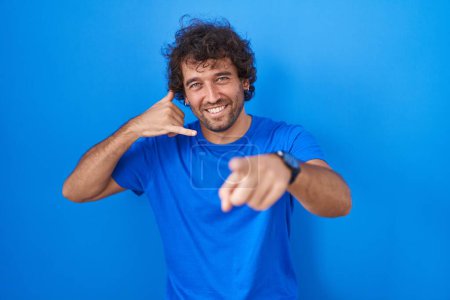 Photo for Hispanic young man standing over blue background smiling doing talking on the telephone gesture and pointing to you. call me. - Royalty Free Image