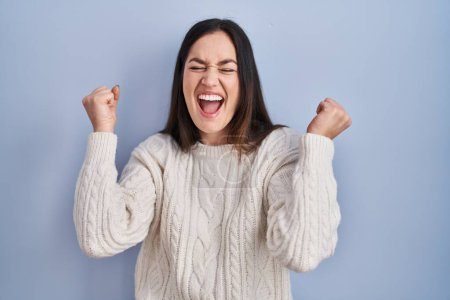 Foto de Young brunette woman standing over blue background celebrating surprised and amazed for success with arms raised and eyes closed. winner concept. - Imagen libre de derechos