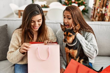 Foto de Dos mujeres sorprenden con bolsa de regalo sentado con perro por árbol de Navidad en casa - Imagen libre de derechos