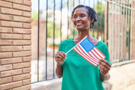 Téléchargez les photos : Moyen Age femme afro-américaine souriante confiante tenant drapeau des États-Unis dans la rue - en image libre de droit