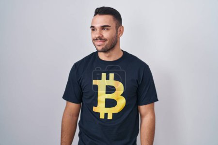 Téléchargez les photos : Jeune homme hispanique portant Bitcoin t-shirt souriant regardant vers le côté et regardant loin en pensant. - en image libre de droit