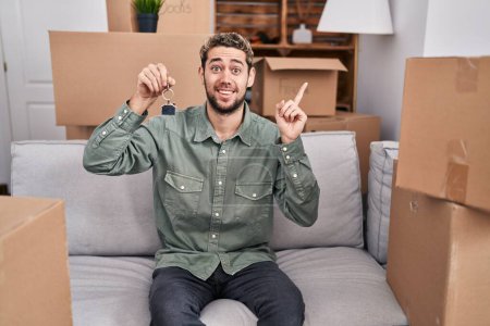 Foto de Hispanic man with beard holding keys of new home smiling with an idea or question pointing finger with happy face, number one - Imagen libre de derechos