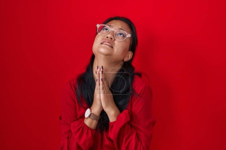 Foto de Mujer joven asiática de pie sobre fondo rojo mendigando y orando con las manos junto con la expresión de esperanza en la cara muy emocional y preocupado. mendicidad. - Imagen libre de derechos