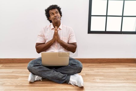 Téléchargez les photos : Homme africain avec les cheveux bouclés à l'aide d'un ordinateur portable assis sur le sol mendier et prier avec les mains ainsi que l'expression de l'espoir sur le visage très émotionnel et inquiet. mendier. - en image libre de droit