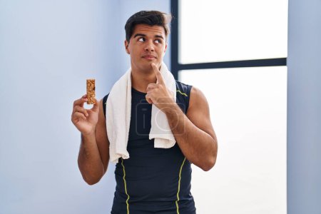 Photo for Hispanic man eating protein bar as healthy energy snack serious face thinking about question with hand on chin, thoughtful about confusing idea - Royalty Free Image