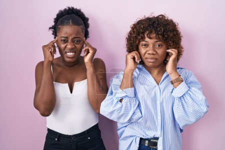 Téléchargez les photos : Two african women standing over pink background covering ears with fingers with annoyed expression for the noise of loud music. deaf concept. - en image libre de droit
