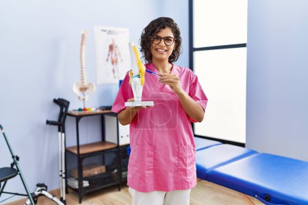 Photo for Young latin woman wearing physiotherapist uniform holding anatomical model of knee at clinic - Royalty Free Image