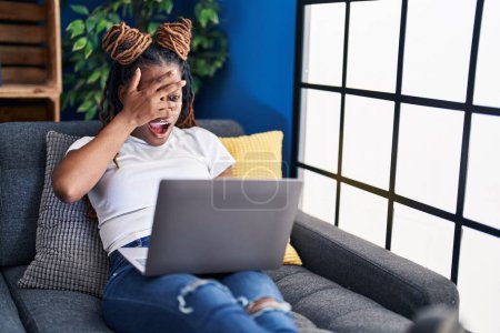 Foto de African woman with braided hair using laptop at home peeking in shock covering face and eyes with hand, looking through fingers afraid - Imagen libre de derechos