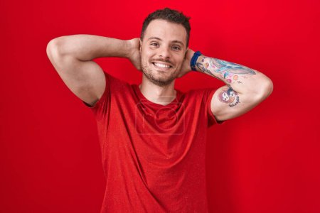 Foto de Joven hombre hispano de pie sobre fondo rojo relajante y estiramiento, brazos y manos detrás de la cabeza y el cuello sonriendo feliz - Imagen libre de derechos