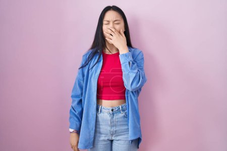 Foto de Joven mujer asiática de pie sobre fondo rosa aburrido bostezo cansado cubriendo la boca con la mano. inquietud y somnolencia. - Imagen libre de derechos