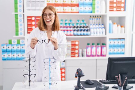 Photo for Young redhead woman working at pharmacy drugstore holding glasses smiling and laughing hard out loud because funny crazy joke. - Royalty Free Image