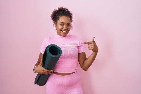 Téléchargez les photos : Young hispanic woman with curly hair holding yoga mat over pink background looking confident with smile on face, pointing oneself with fingers proud and happy. - en image libre de droit