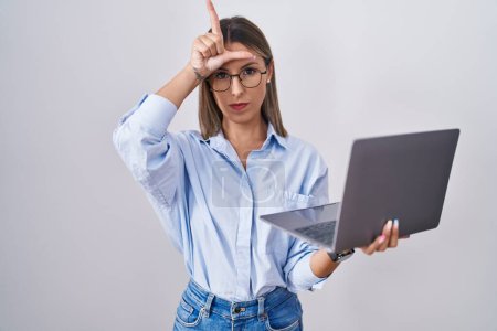 Foto de Young woman working using computer laptop making fun of people with fingers on forehead doing loser gesture mocking and insulting. - Imagen libre de derechos