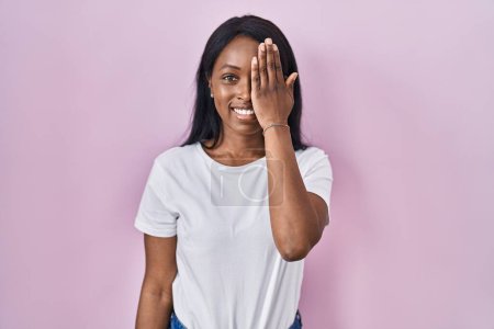 Foto de Mujer joven africana con camiseta blanca casual que cubre un ojo con la mano, sonrisa confiada en la cara y emoción sorpresa. - Imagen libre de derechos