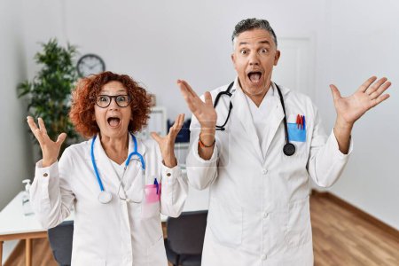 Foto de Dos médicos de mediana edad en la clínica médica celebrando la victoria con sonrisa feliz y expresión ganadora con las manos levantadas - Imagen libre de derechos