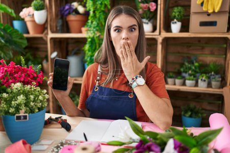 Téléchargez les photos : Hispanic young woman working at florist shop showing smartphone screen covering mouth with hand, shocked and afraid for mistake. surprised expression - en image libre de droit